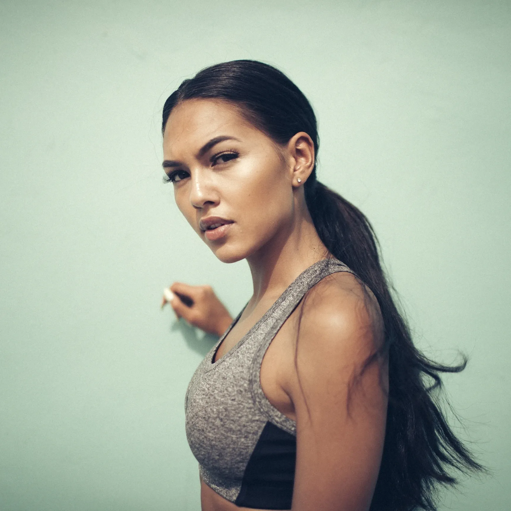 Person mit Pferdeschwanz in grauem Sporttop vor türkisfarbener Wand, blickt ernst zur Kamera