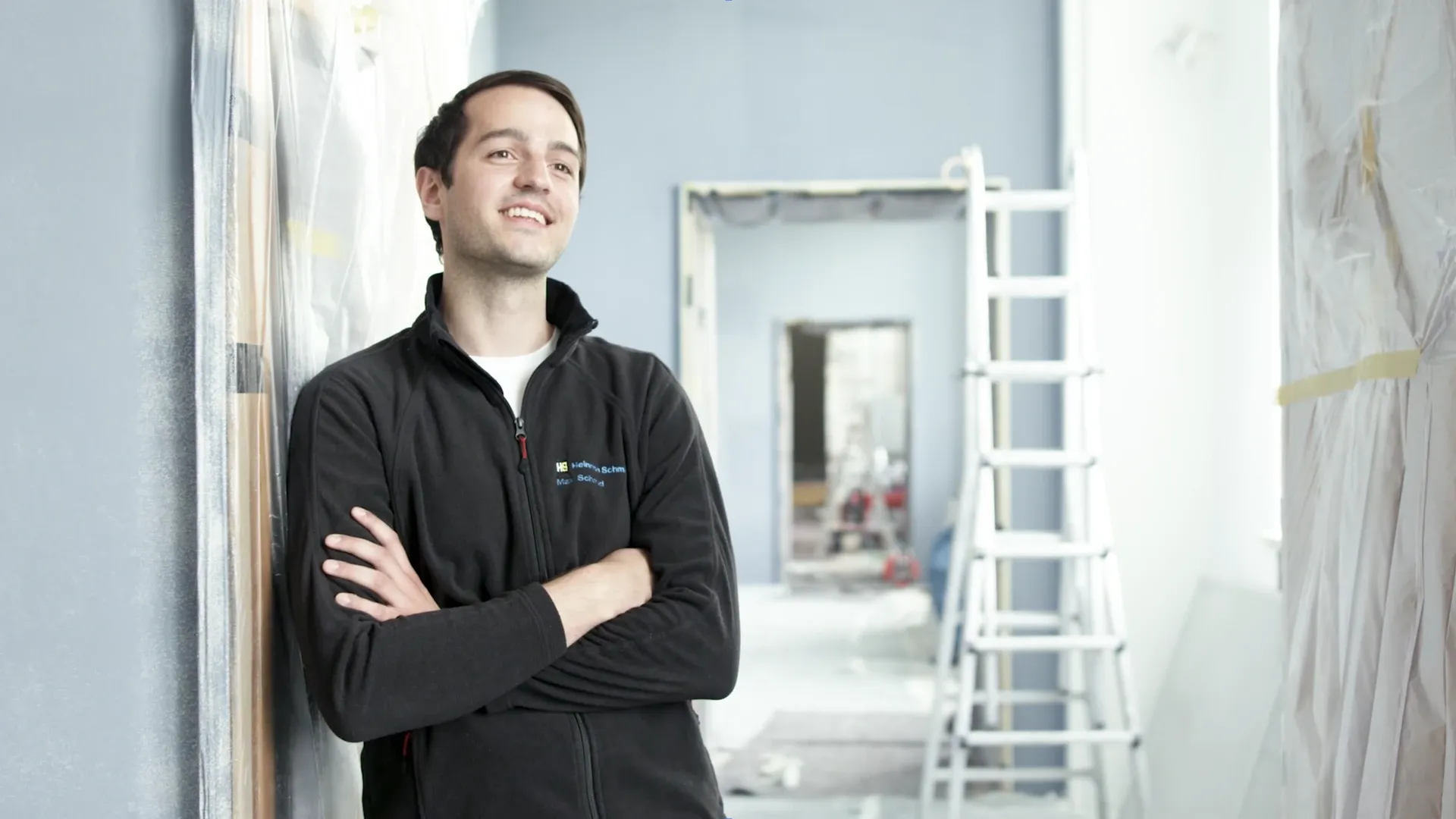 Lächelnder Mann mit verschränkten Armen lehnt an einer Wand, im Hintergrund Leiter und abgedeckte Flächen