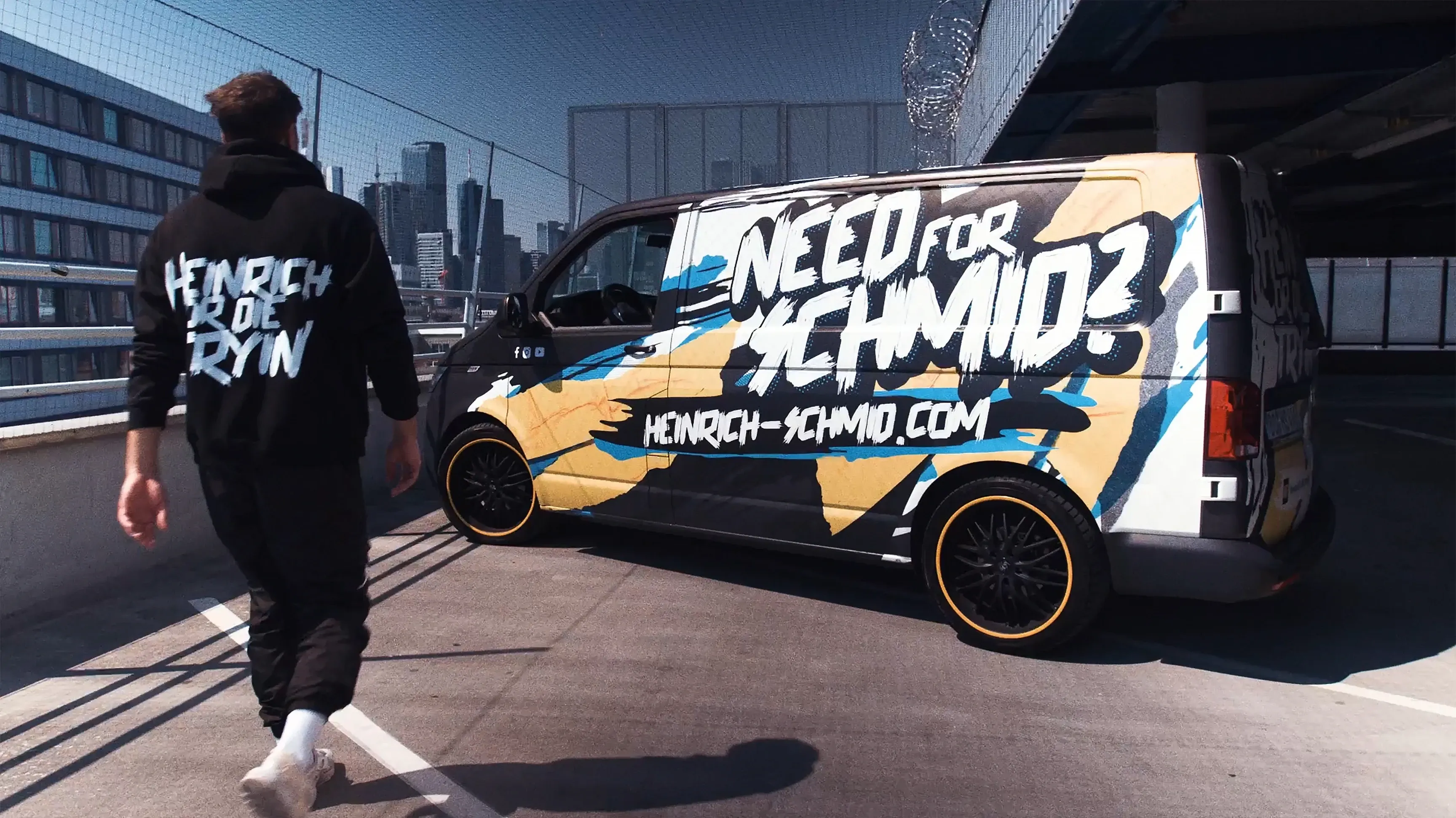 Mann auf Parkdeck geht auf bunt folierten Van mit der Aufschrift "NEED FOR SCHMID?" zu