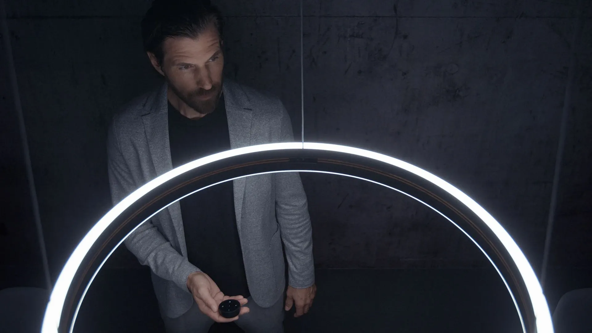 Mann unter modernem, ringförmigem LED-Licht, steuert die Leuchte mit einer Fernbedienung – stilvolle Beleuchtung in dunkler Umgebung.