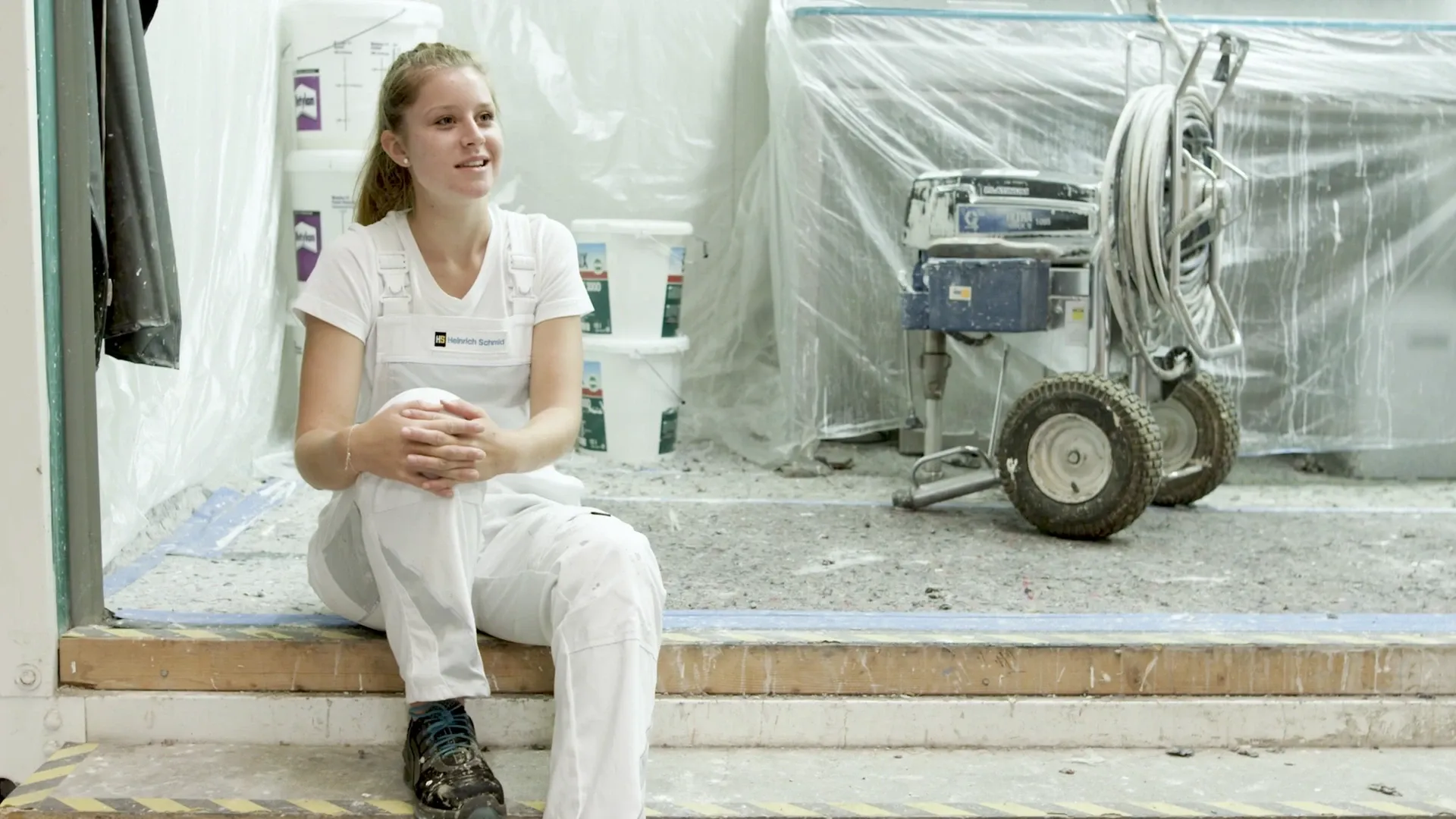 Person in weißer Malerkleidung sitzt auf einer Stufe vor abgedeckten Wänden, neben Farbeimern und Farbspritzgerät