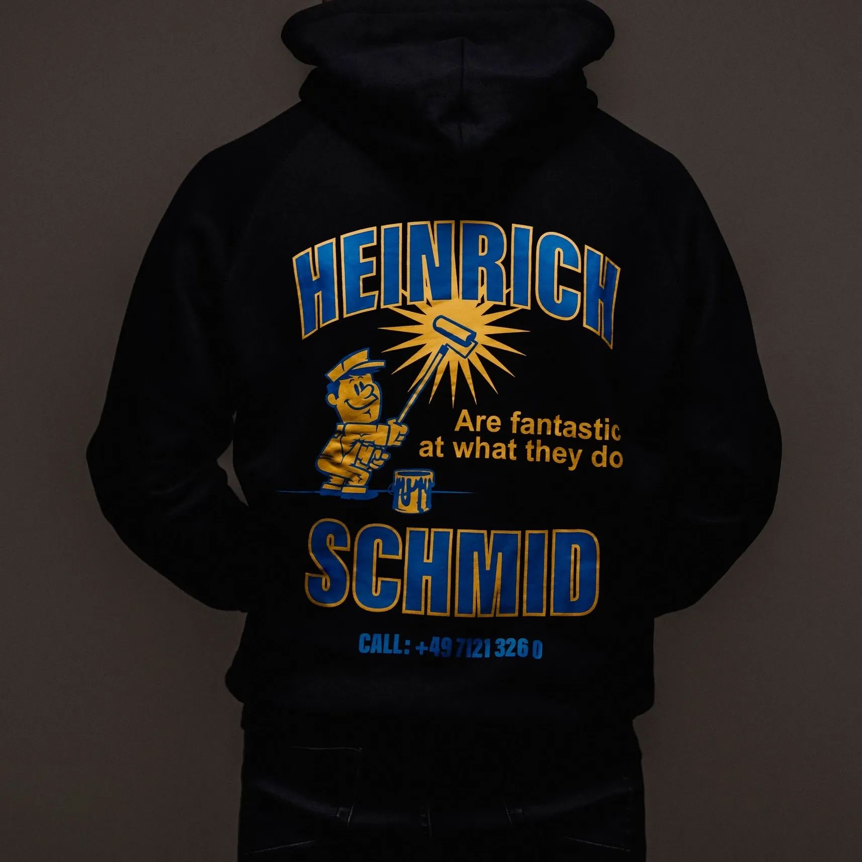 Person von hinten in schwarzem Hoodie mit blau-gelbem „Heinrich Schmid“-Aufdruck und Cartoon-Handwerker