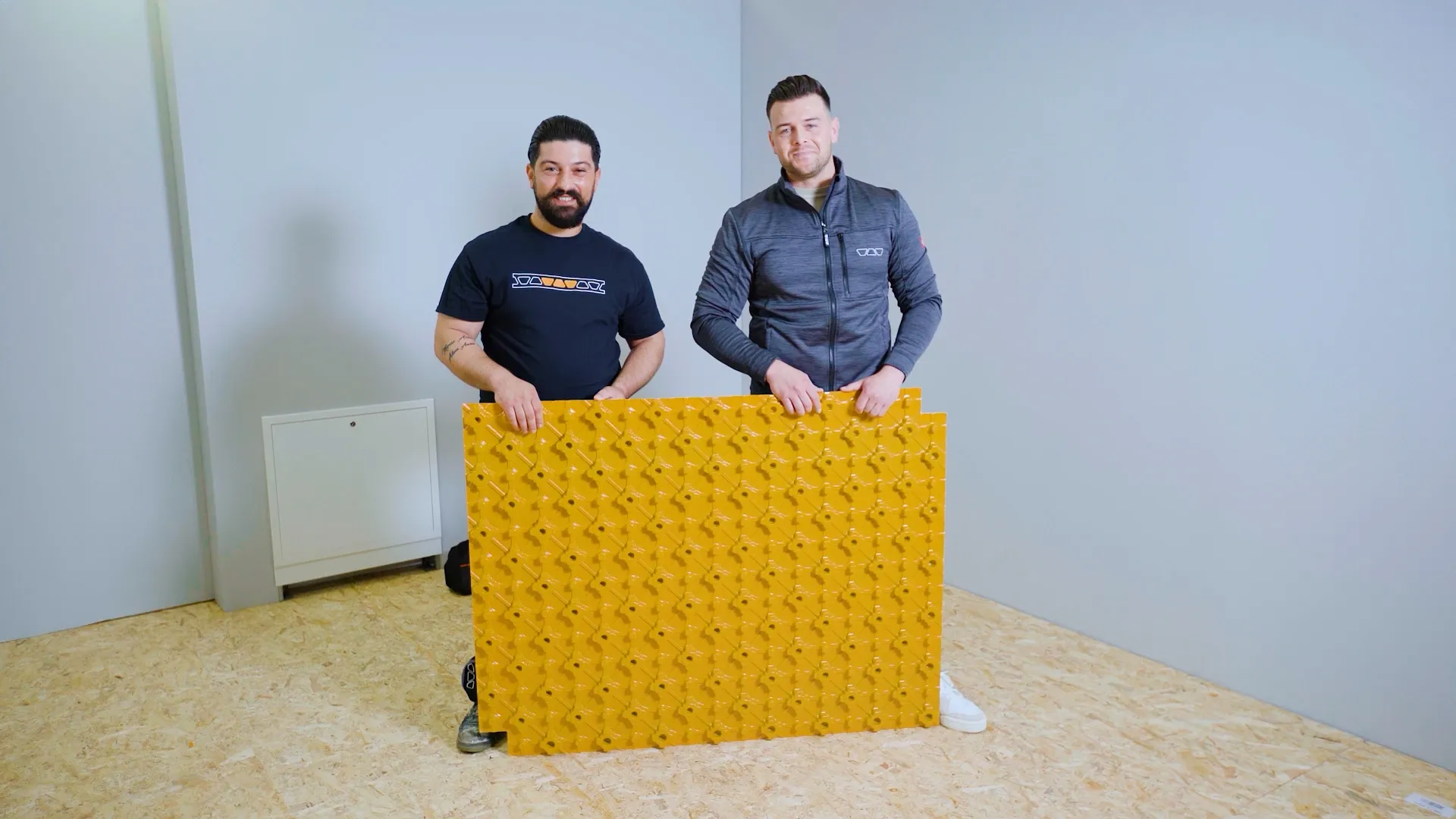 Zwei Männer halten eine große gelbe Noppenplatte in einem leeren Raum