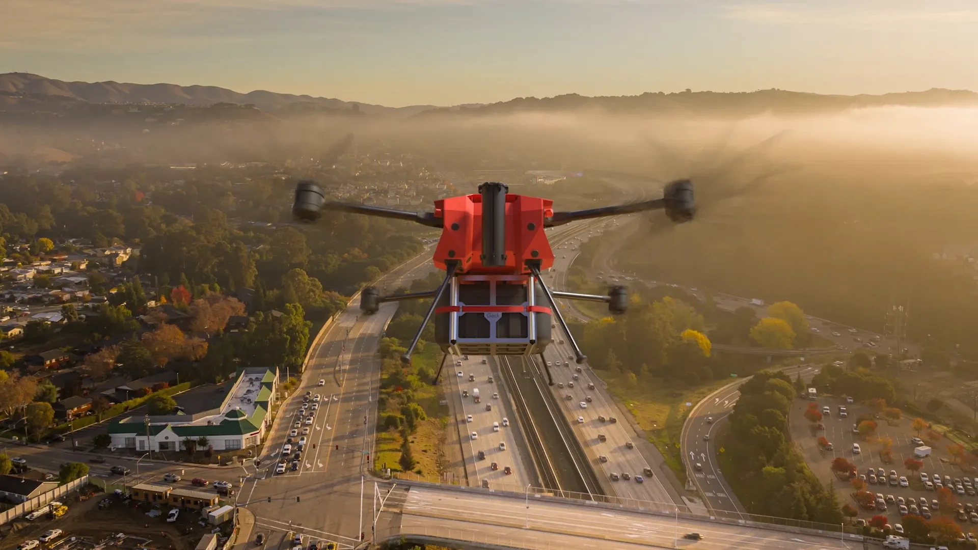 Rote Multikopter-Drohne fliegt über einer Autobahnbrücke bei Nebel, Stadt und Hügel im Hintergrund