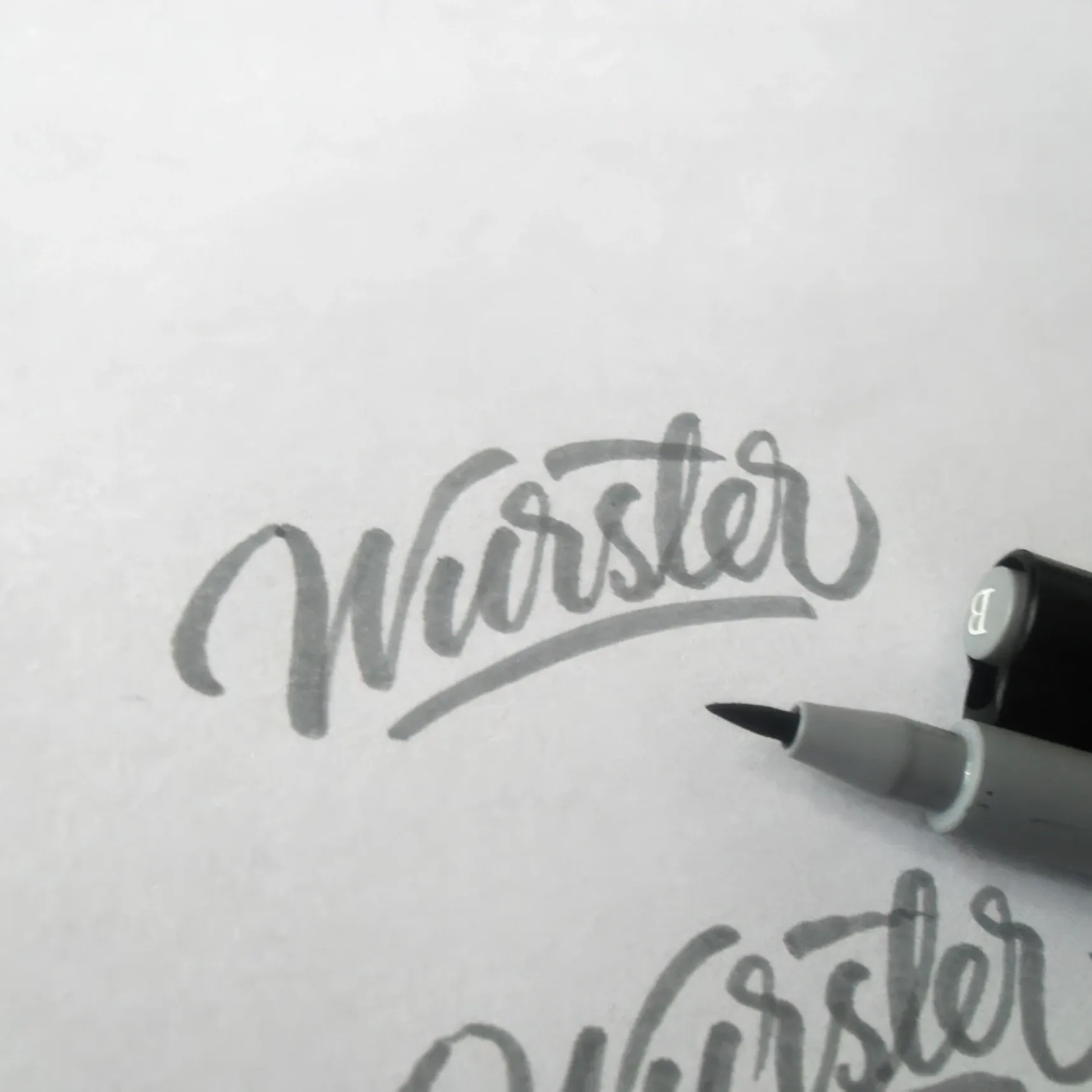 Grauer Brushpen neben dem Handlettering „Würster“ auf weißem Papier