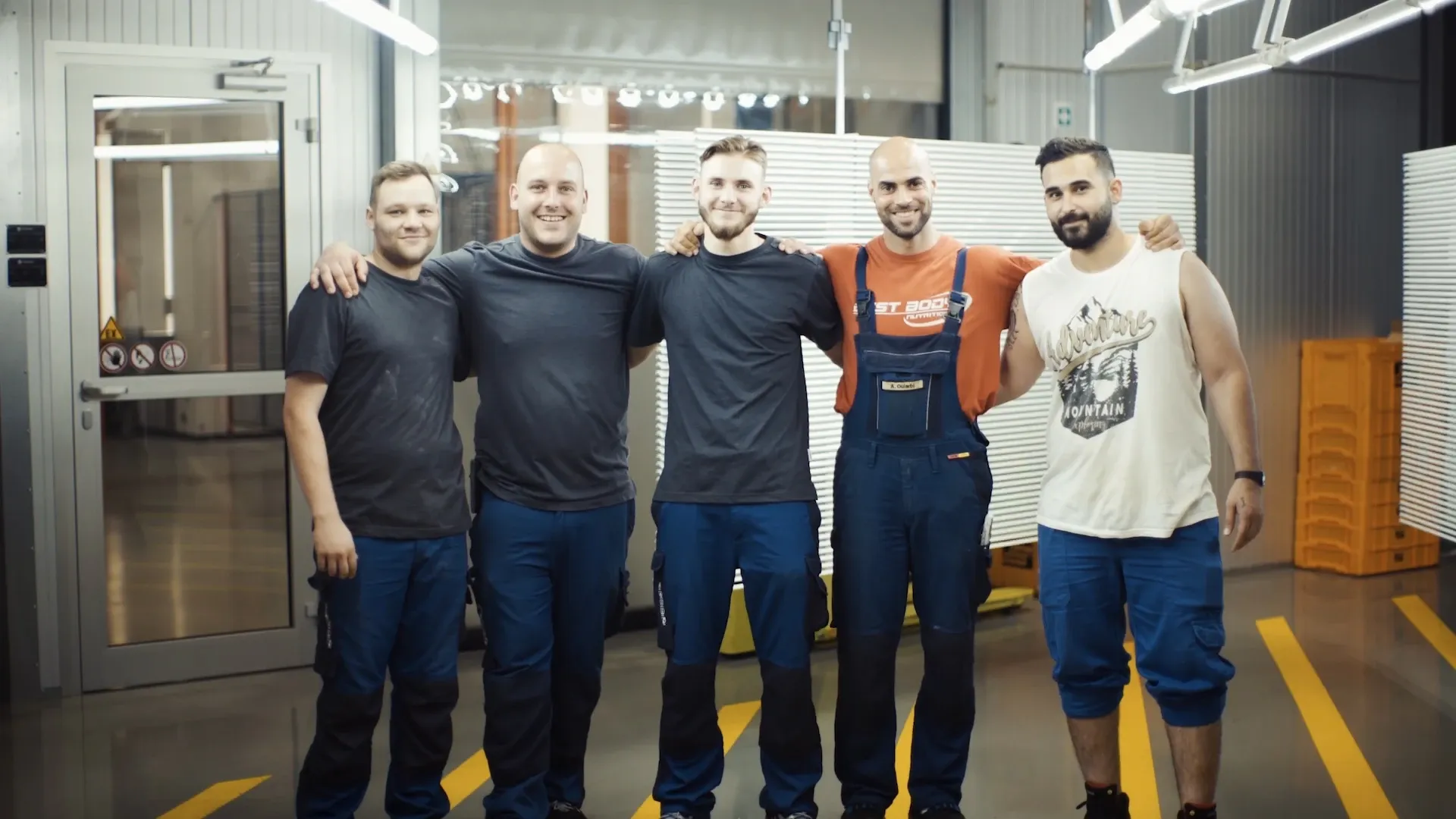 Fünf Männer in Arbeitskleidung posieren Arm in Arm lächelnd in einer Werkhalle