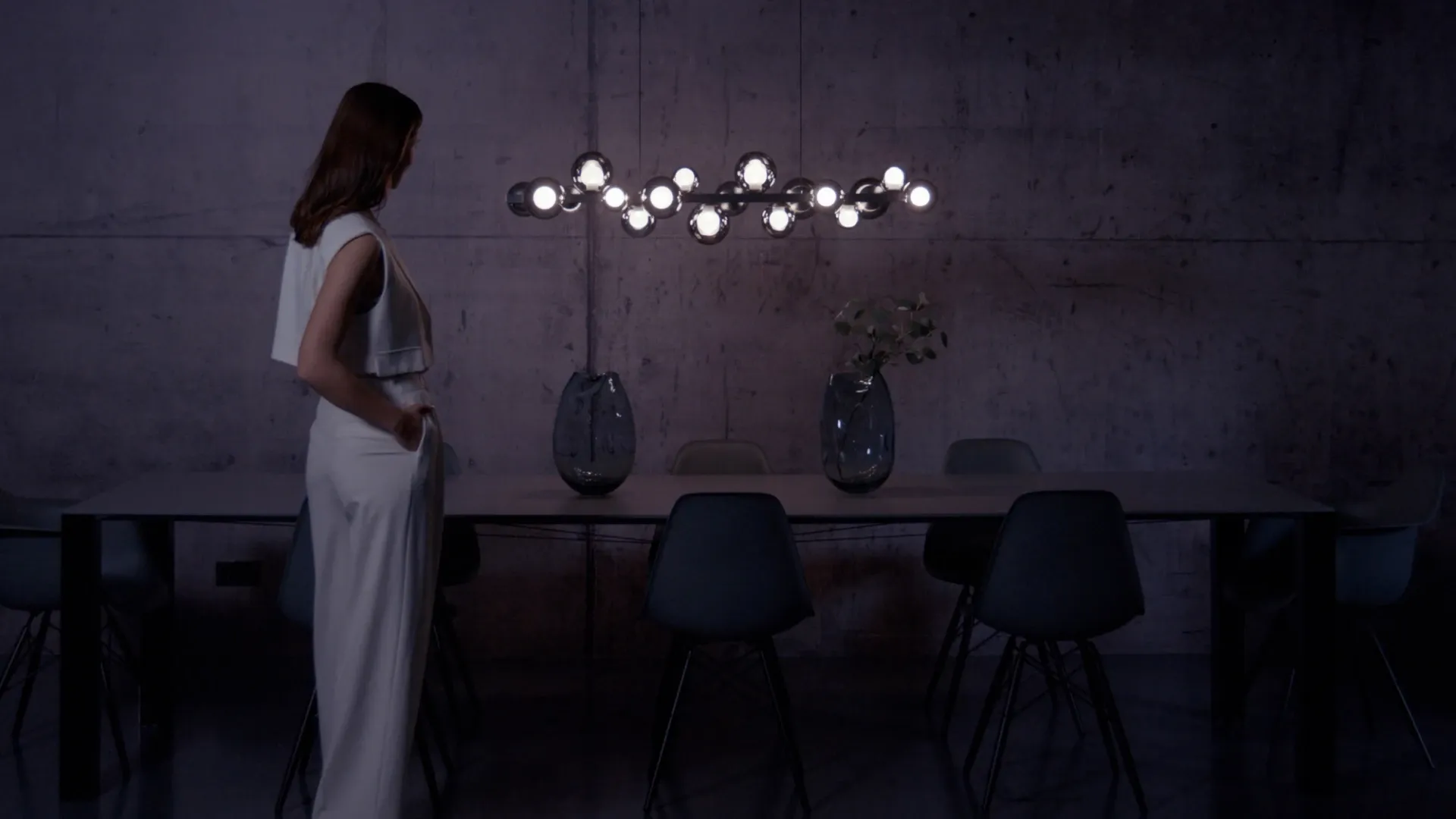 Person in heller Kleidung steht neben einem Esstisch mit moderner Hängelampe in einem dunklen Raum mit Betonwand.