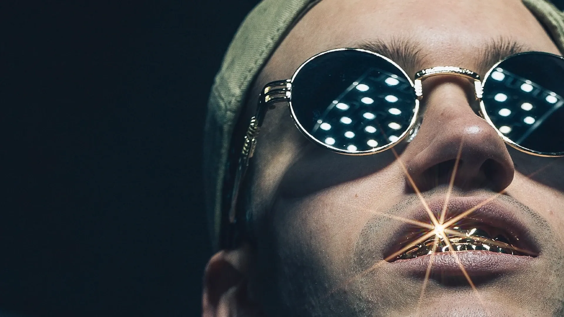 Nahaufnahme einer Person mit Mütze, runden Sonnenbrillen und funkelnden goldenen Grillz