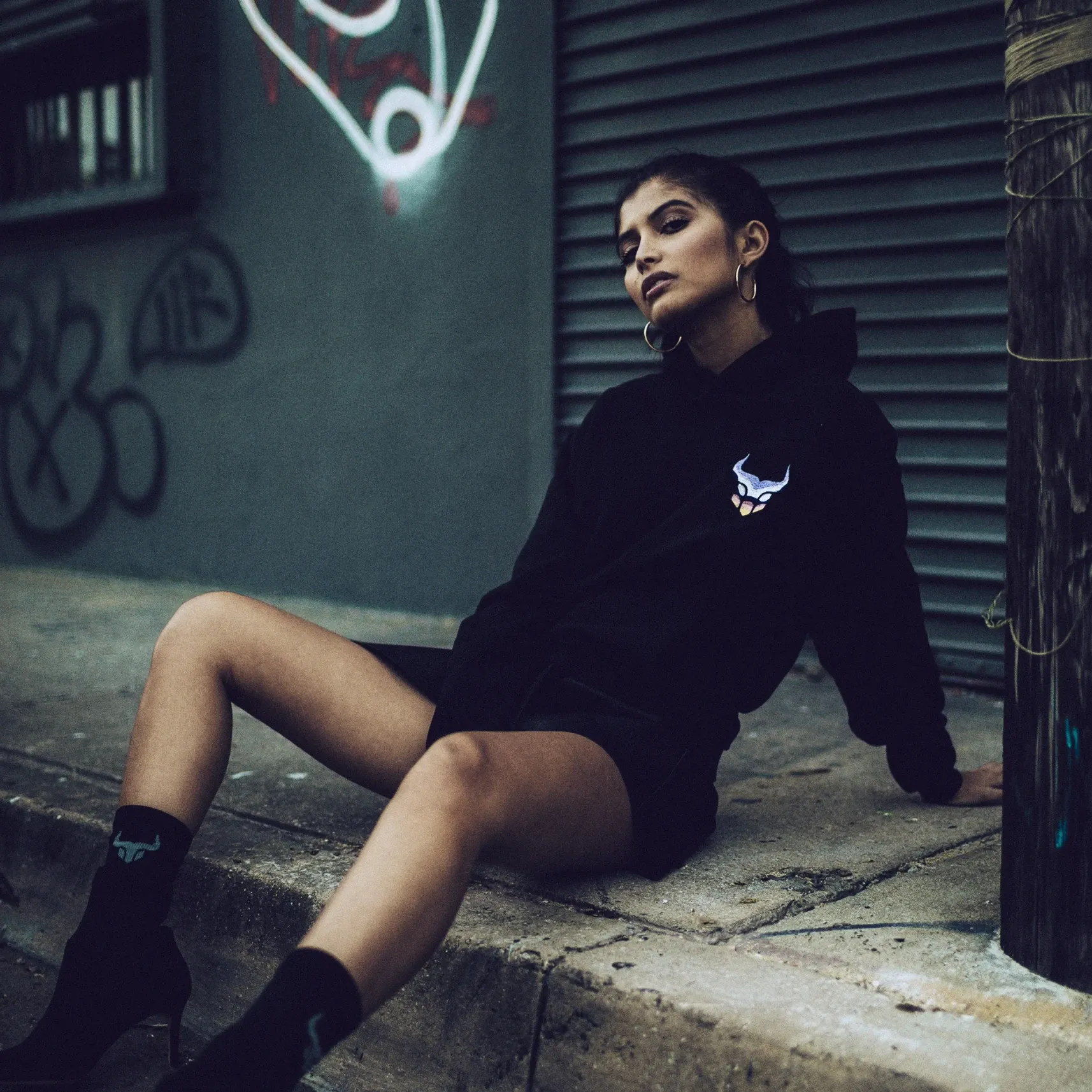 Frau in schwarzem Hoodie sitzt auf dem Bordstein vor einem Rolltor, Graffiti an der Wand