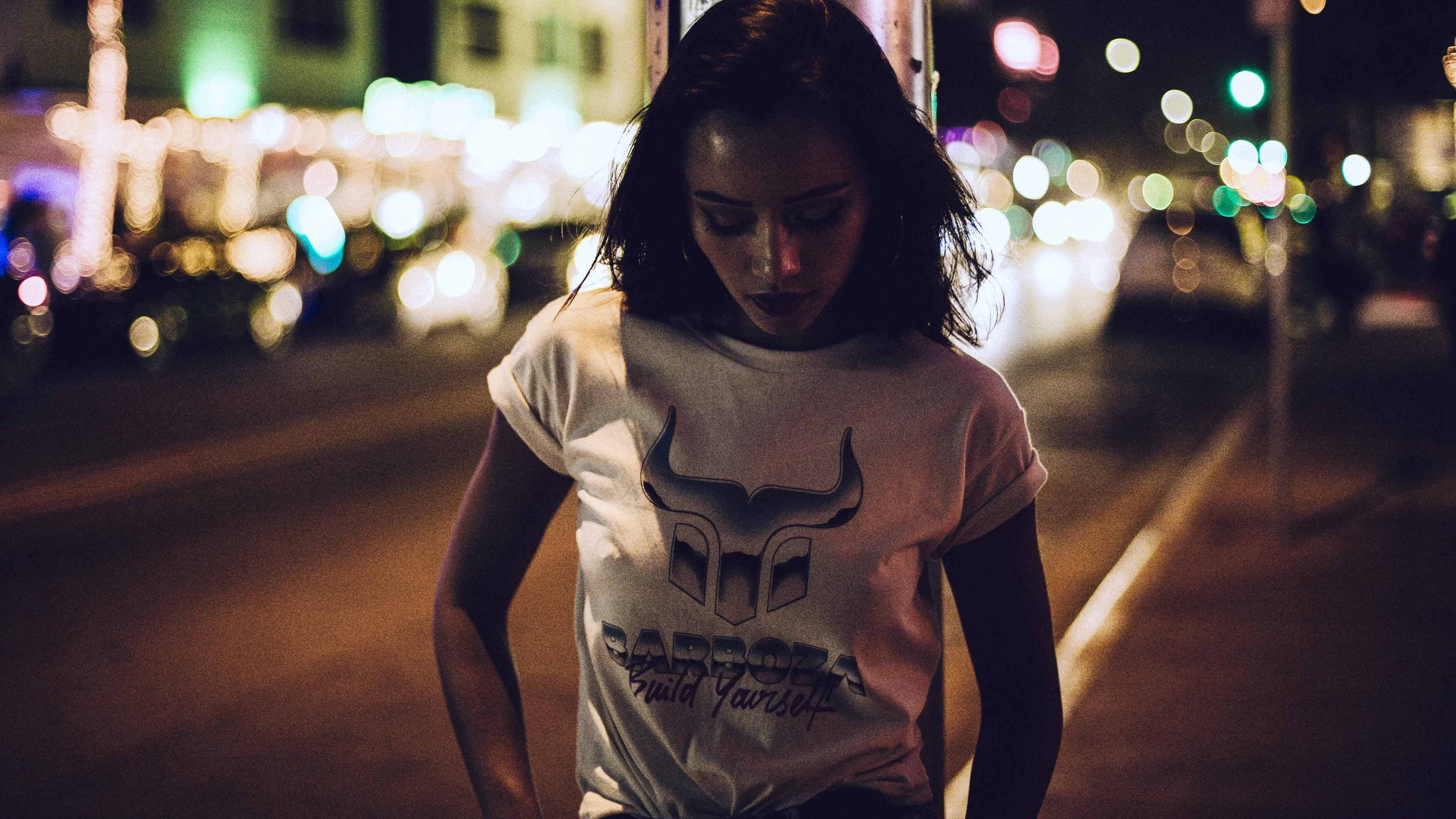 Person mit weißem T‑Shirt mit Stier-Logo auf nächtlicher Straße, unscharfe Lichter im Hintergrund