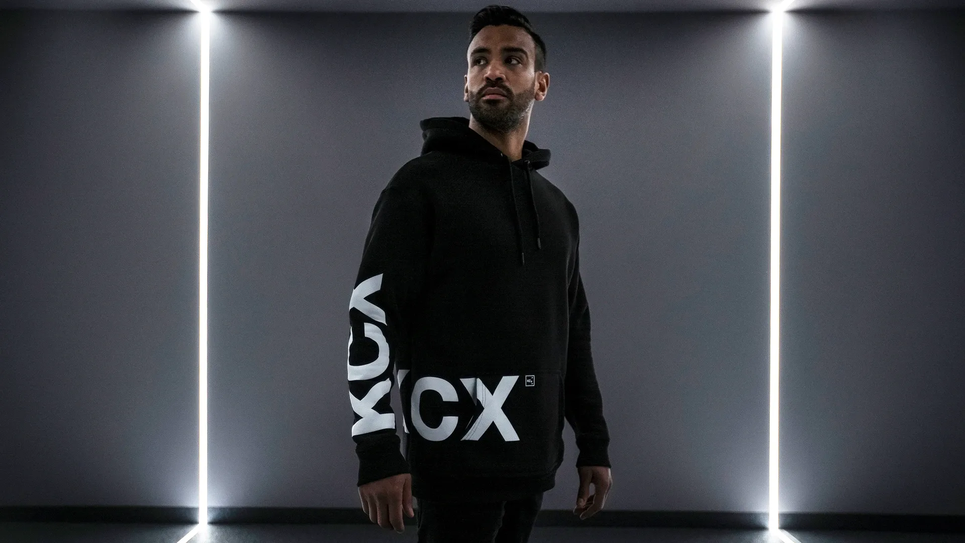 Person in schwarzem Hoodie mit weißem Aufdruck steht in einem dunklen Raum mit vertikalen Lichtstreifen im Hintergrund.