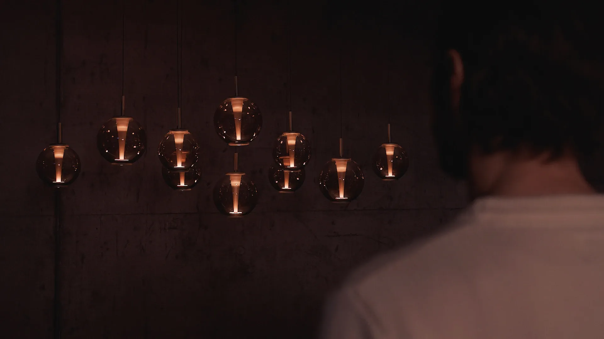 Mehrere kugelförmige Pendelleuchten mit warmem Licht hängen vor einer dunklen Wand, im Vordergrund ist eine Person von hinten zu sehen.
