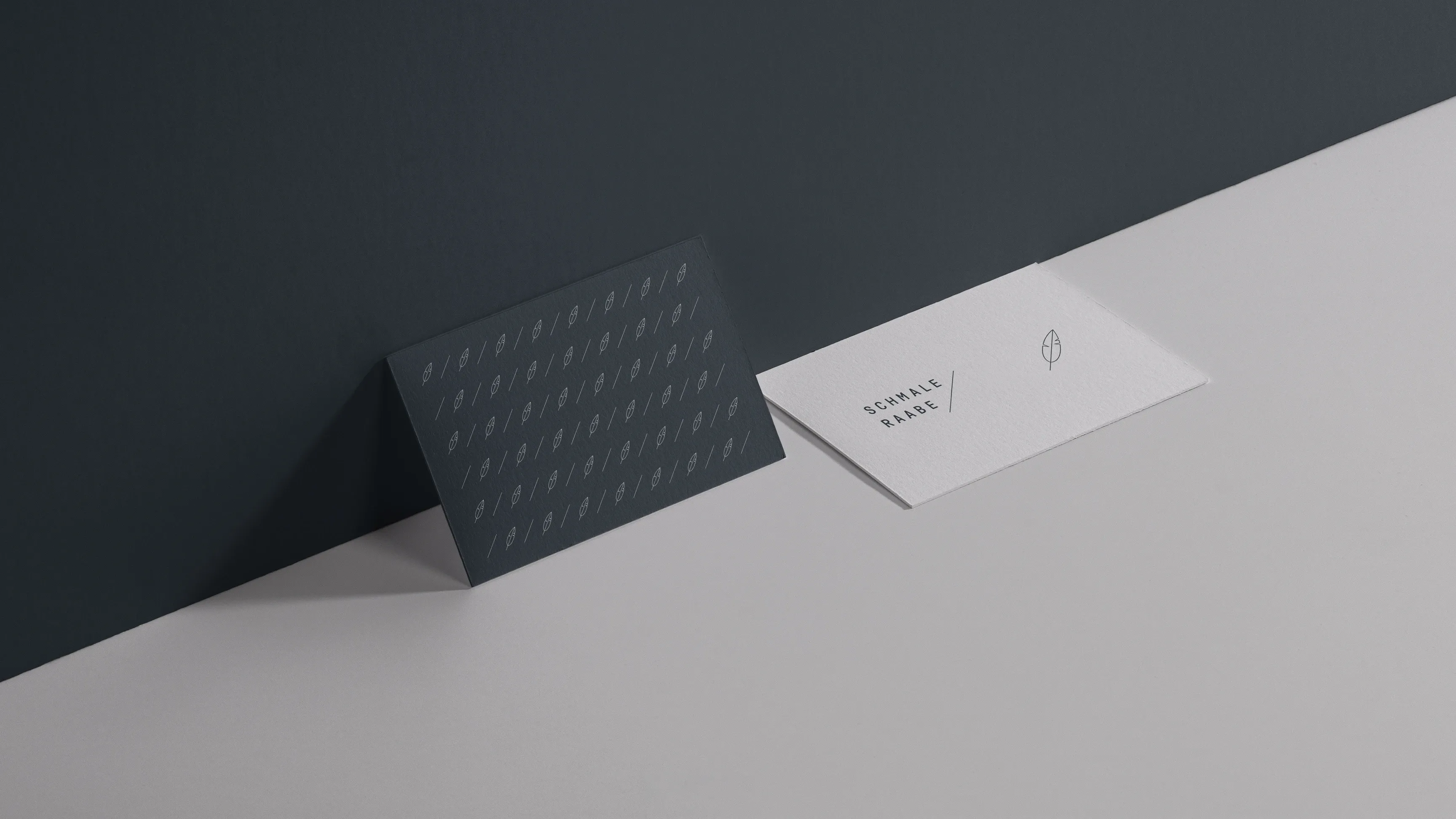 Zwei minimalistische Visitenkarten: eine dunkelgraue mit Muster lehnt an der Wand, eine weiße mit Logo liegt auf grauer Oberfläche