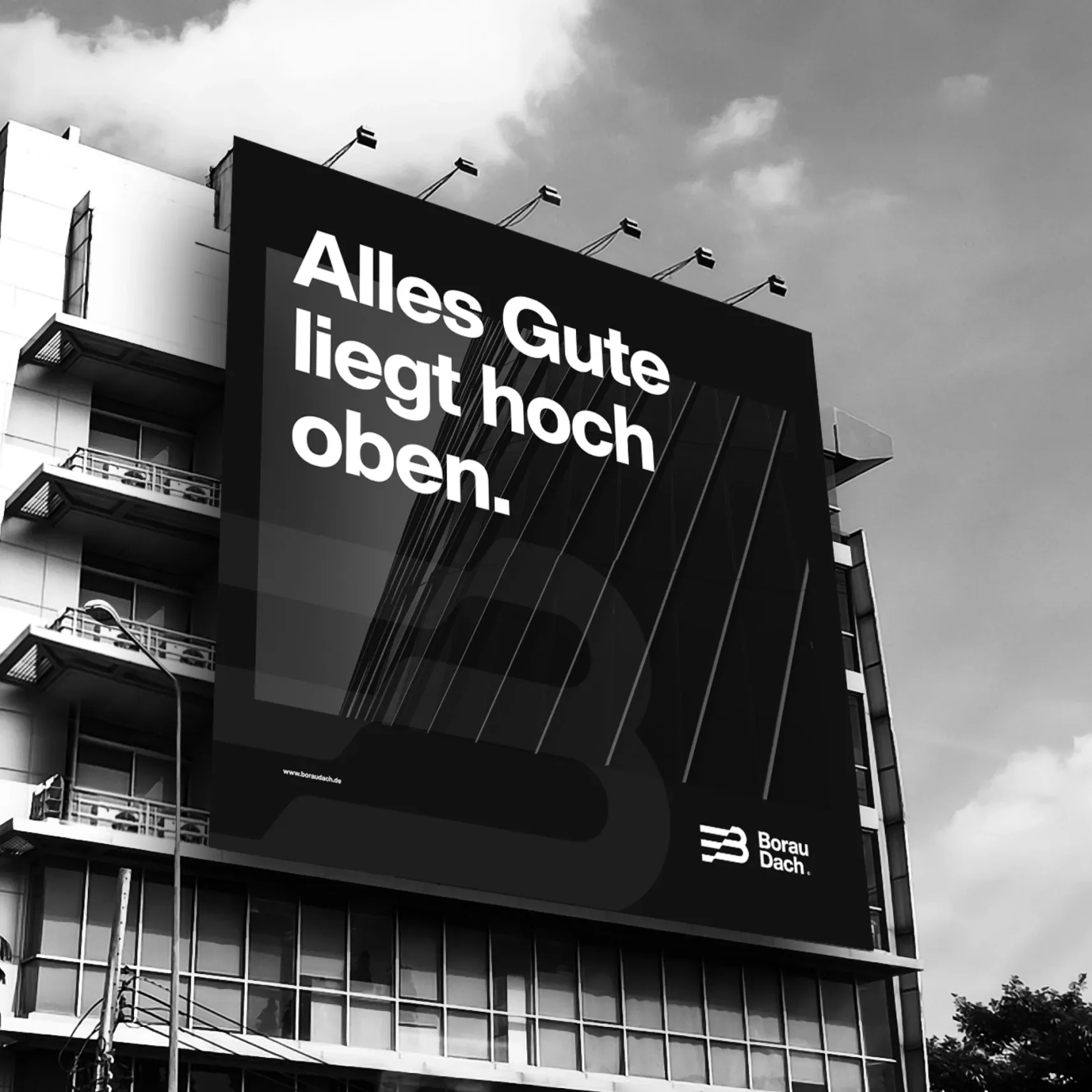 Schwarzweißfoto eines großen Werbebanners an einer Fassade mit dem Text 'Alles Gute liegt hoch oben' und Borau-Dach-Logo