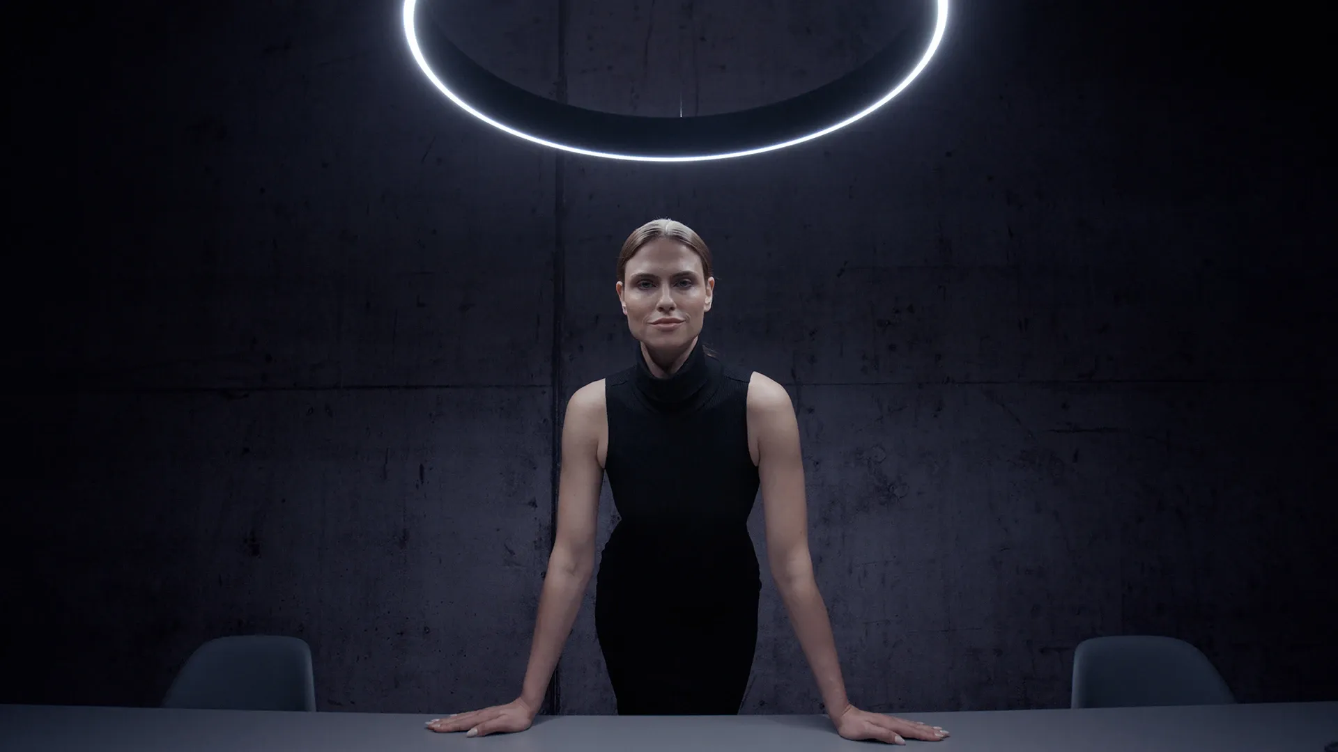 Porträtaufnahme in modernem, minimalistischem Raum – Frau steht unter einem leuchtenden Ringlicht über einem Tisch.