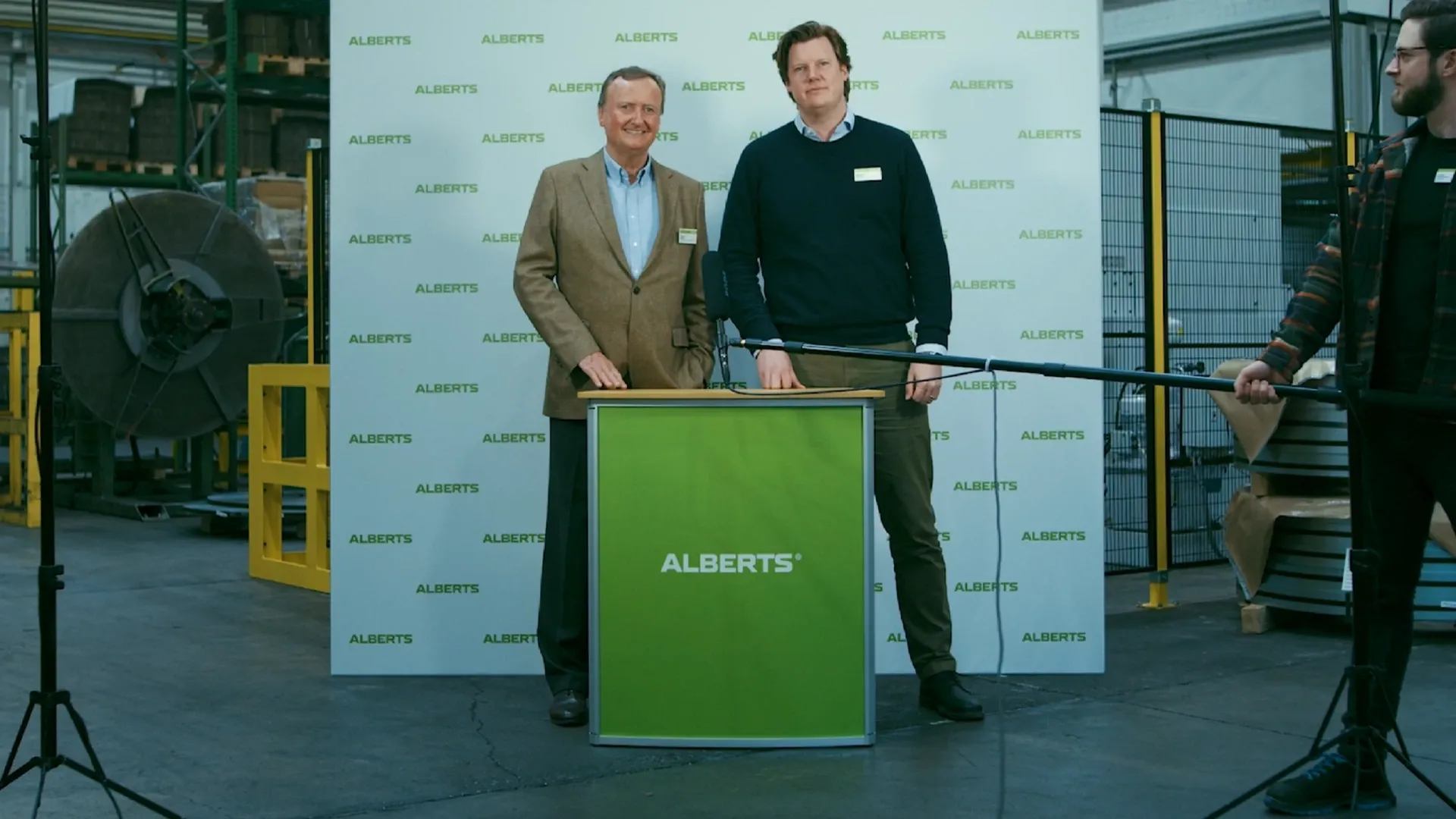 Zwei Männer hinter grünem Podest mit ALBERTS-Logo vor Logo-Hintergrund in einer Industriehalle, rechts hält jemand eine Tonangel