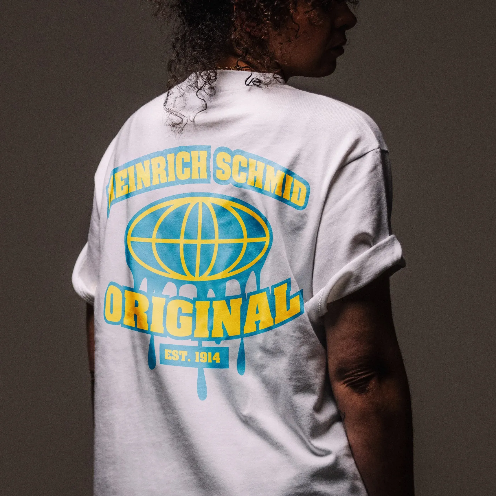 Person von hinten in weißem T‑Shirt mit blau‑gelbem Aufdruck „HEINRICH SCHMID ORIGINAL“ und Globusgrafik