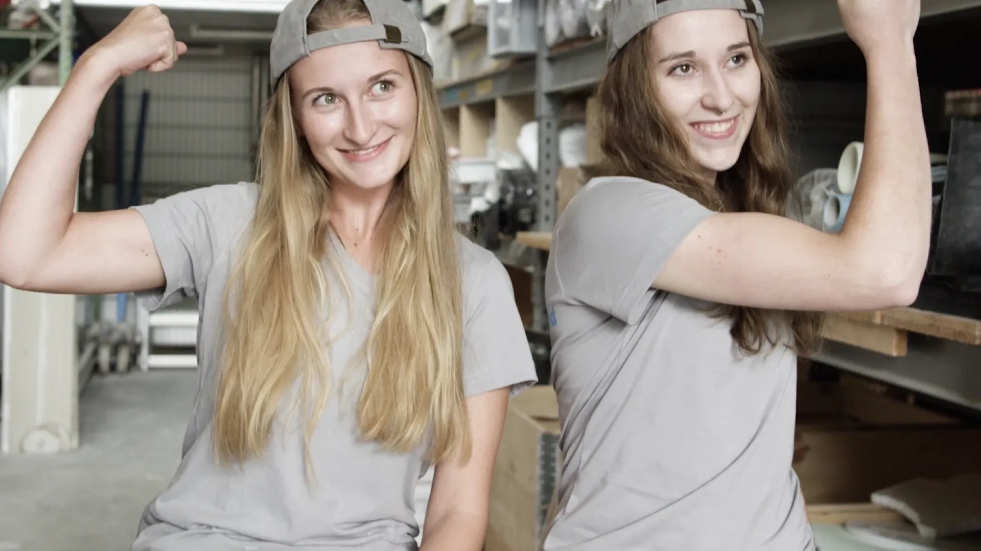 Zwei Personen in grauen T‑Shirts und rückwärts getragenen Caps zeigen lachend ihre Muskeln in einer Werkstatt