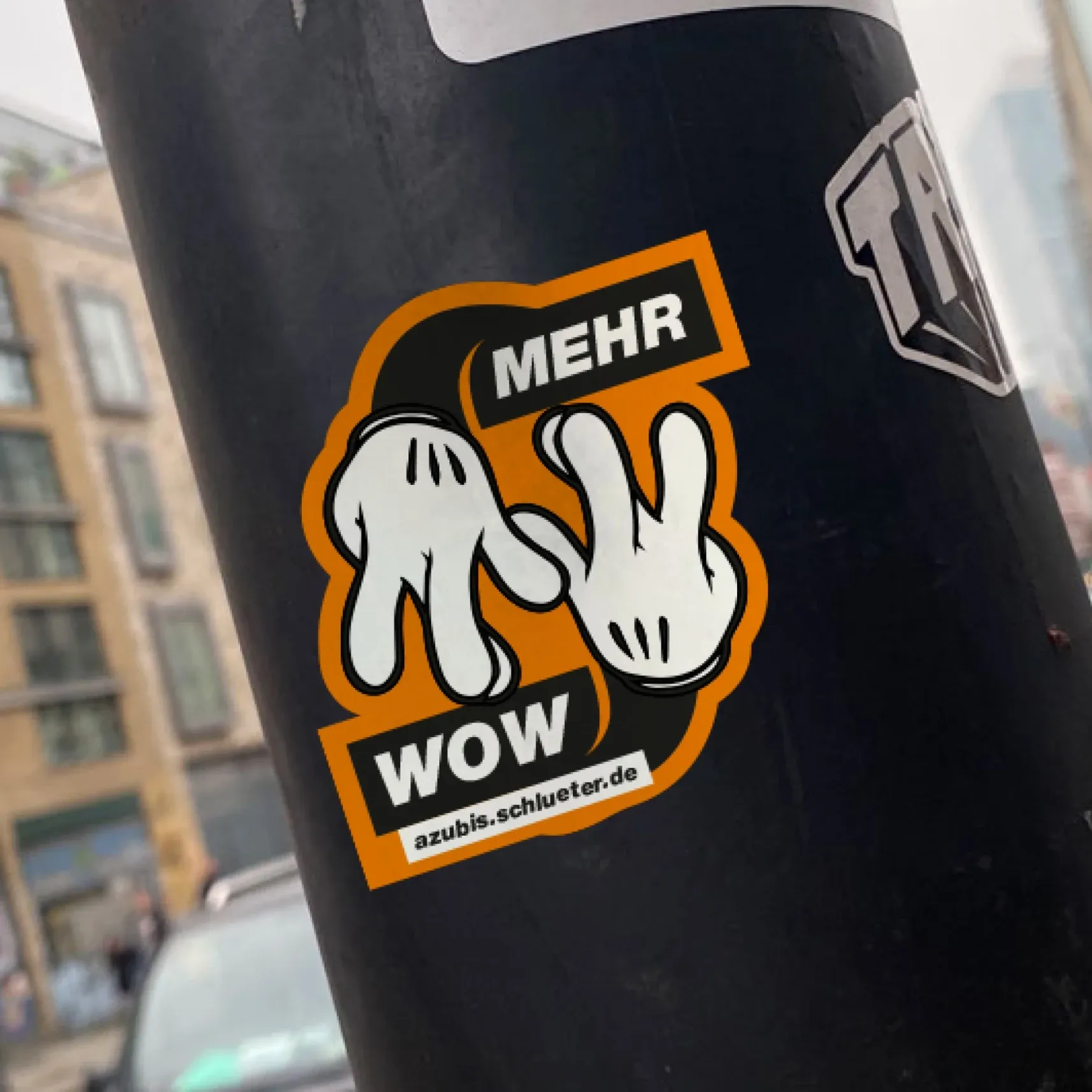 Aufkleber mit weißen Cartoon-Handschuhen und der Aufschrift „MEHR WOW“ an einem schwarzen Pfosten