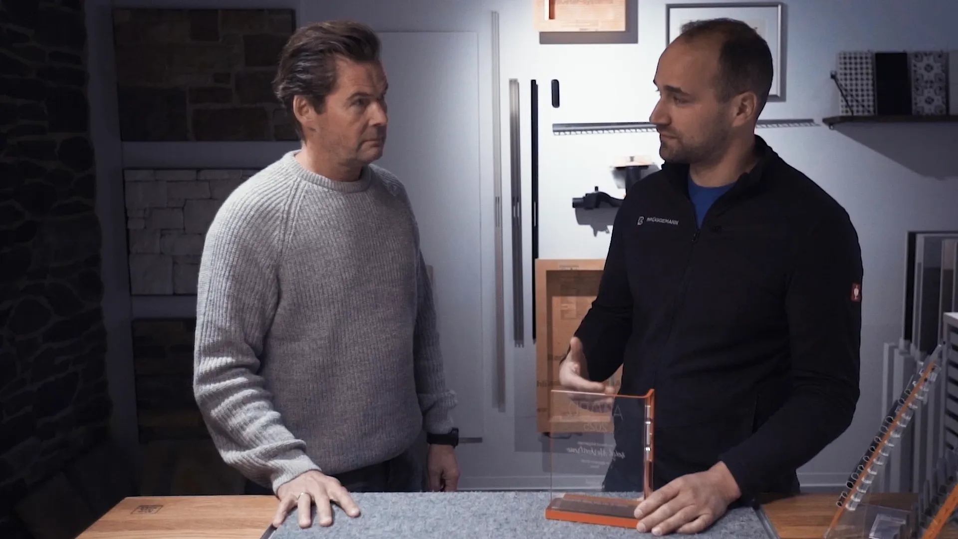 Zwei Männer an einer Arbeitsplatte, der rechte zeigt eine transparente Acryltafel, Materialmuster im Hintergrund