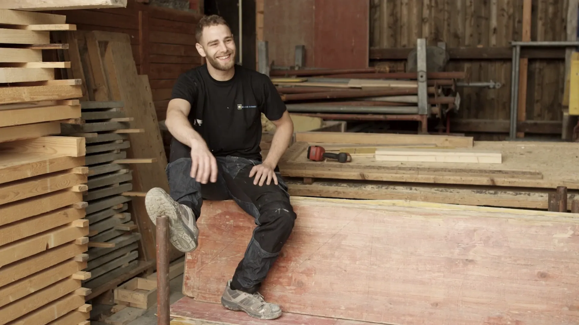 Lächelnder Mann in Arbeitskleidung sitzt in einer Holzwerkstatt, umgeben von Brettern und einem roten Akkuschrauber