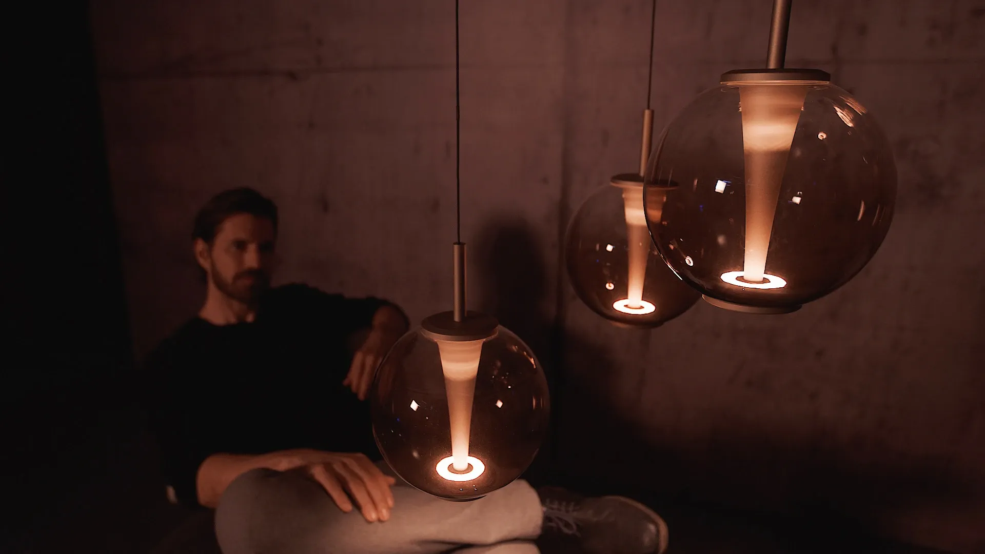 Drei kugelförmige Pendelleuchten leuchten warm in einem dunklen Raum, im Hintergrund sitzt eine Person auf einem Stuhl.