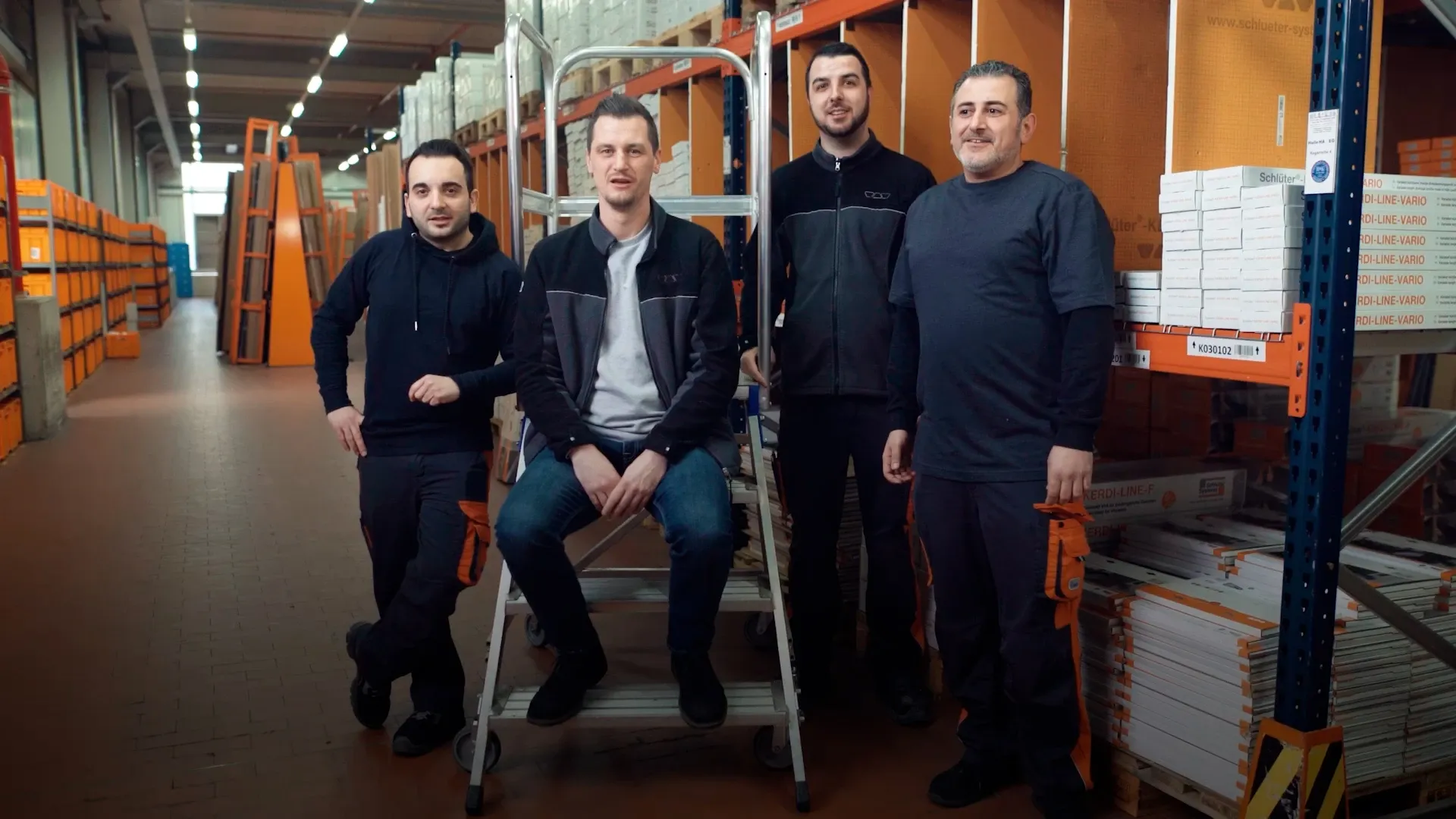 Vier Personen in einem Lager zwischen Regalen und Kartons, eine sitzt auf einer Rollleiter