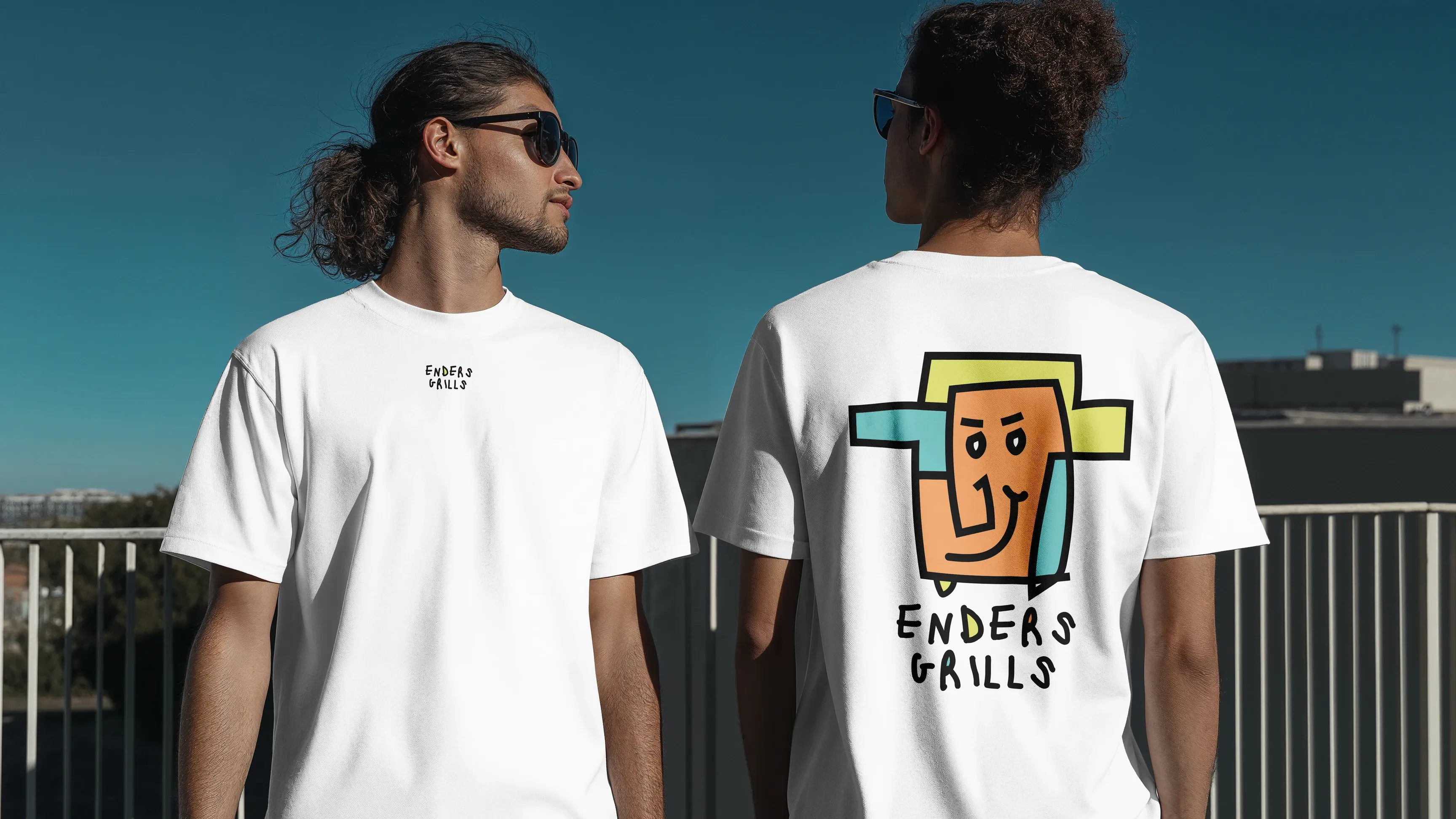 Zwei Personen in weißen T‑Shirts mit „Enders Grills“-Logo, eine zeigt die Rückseite mit buntem Gesichtsmotiv, beide mit Sonnenbrillen auf einem Dach