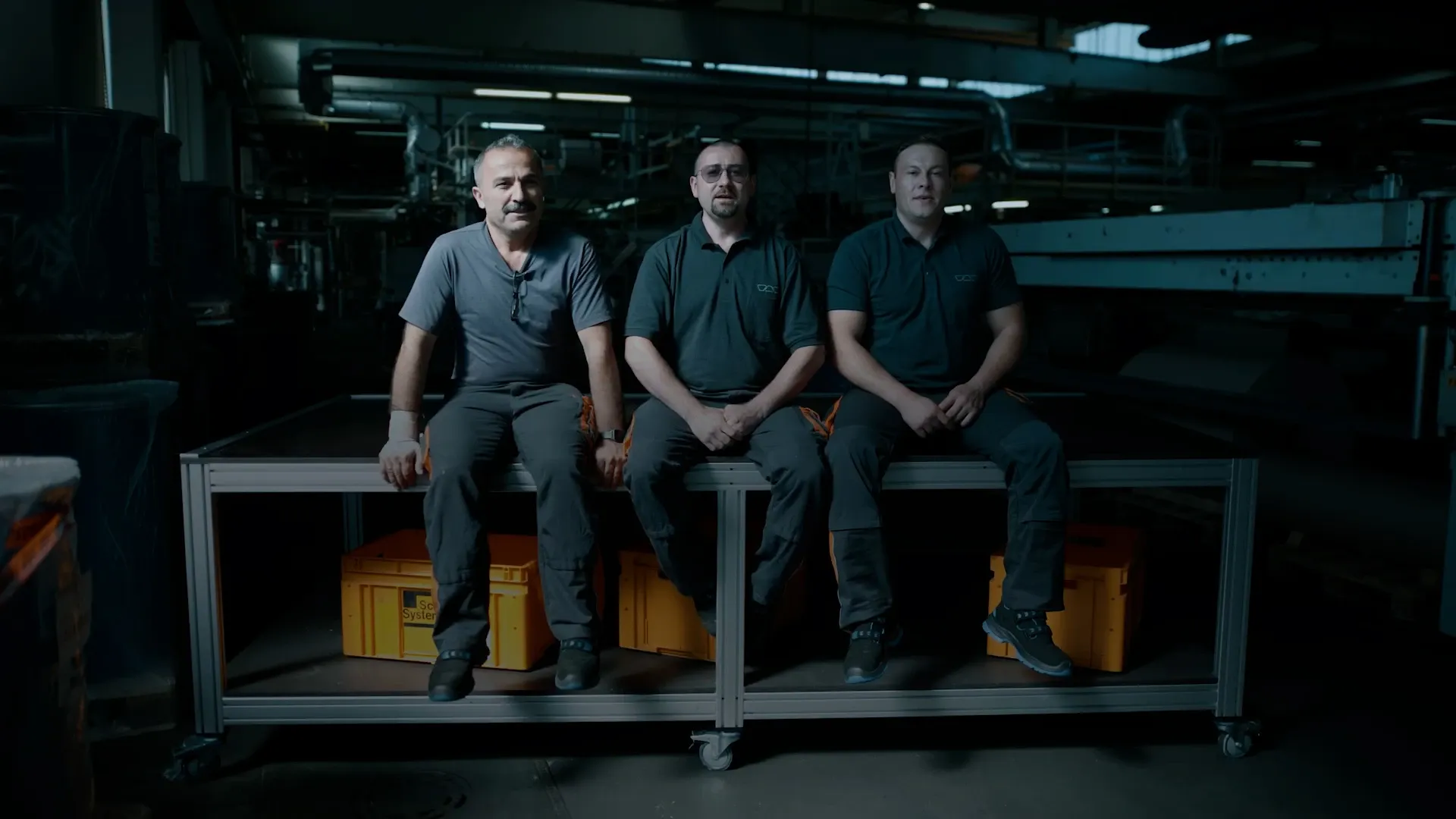 Drei Männer in Arbeitskleidung sitzen auf einer Werkbank in einer Fabrikhalle, gelbe Kisten darunter