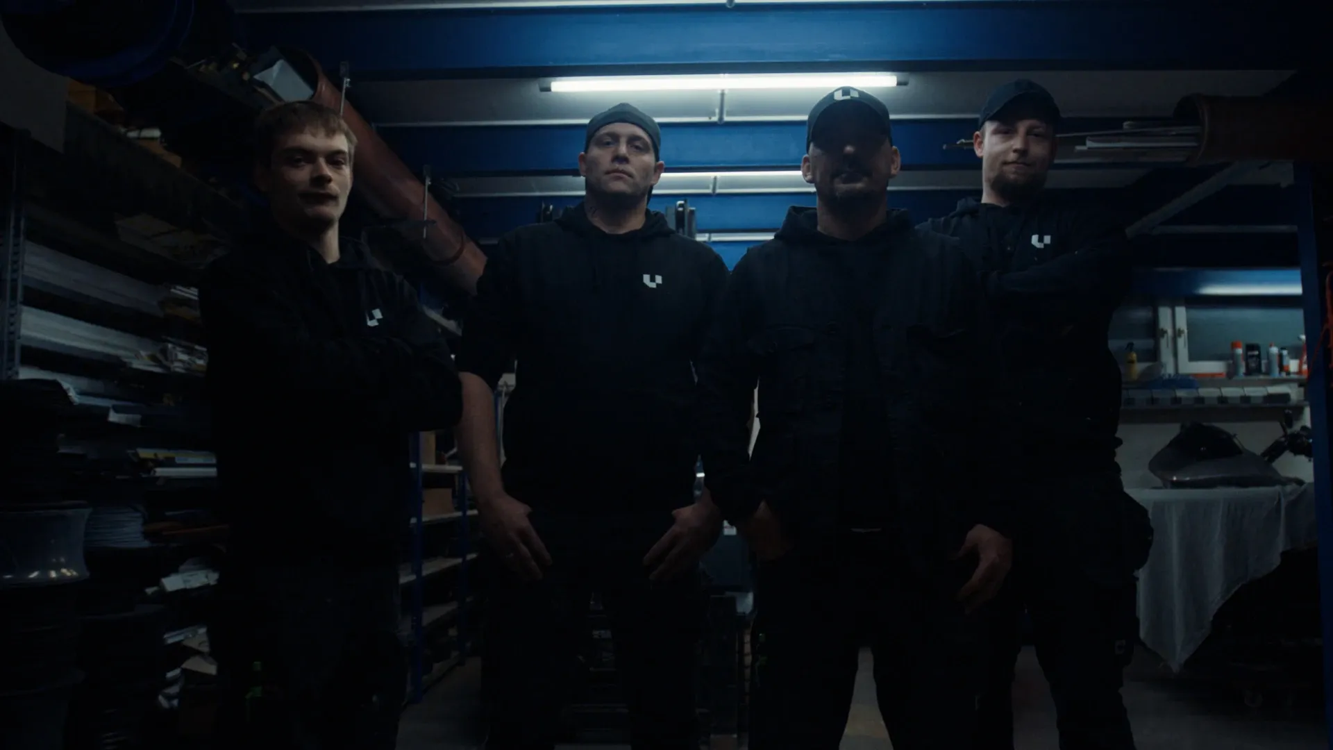 Vier Männer in schwarzer Kleidung stehen in einer dunkel beleuchteten Werkstatt