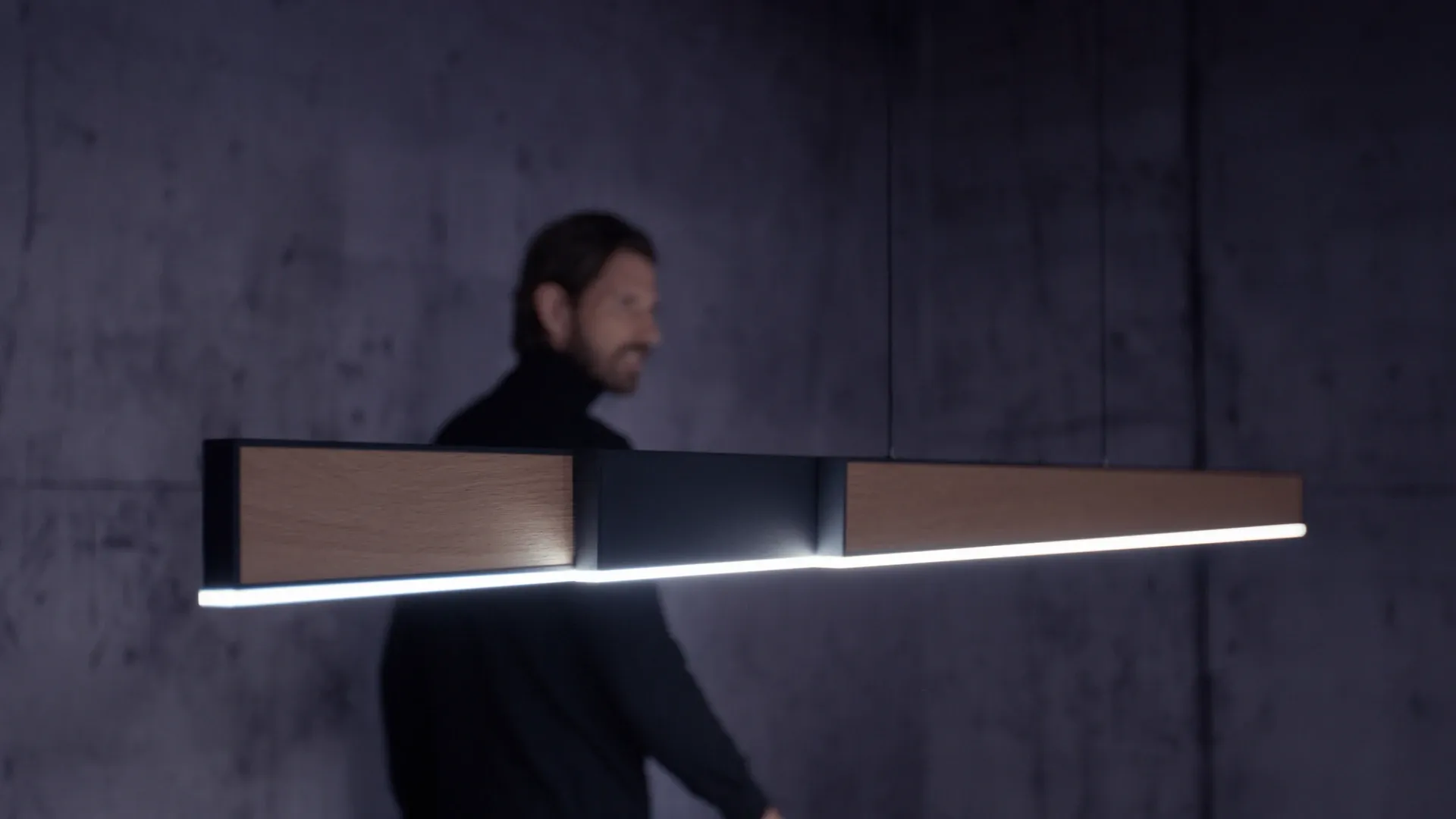 Lineare Pendelleuchte mit Holzpaneelen und LED-Lichtleiste vor dunkler Betonwand, unscharfe Person im Hintergrund