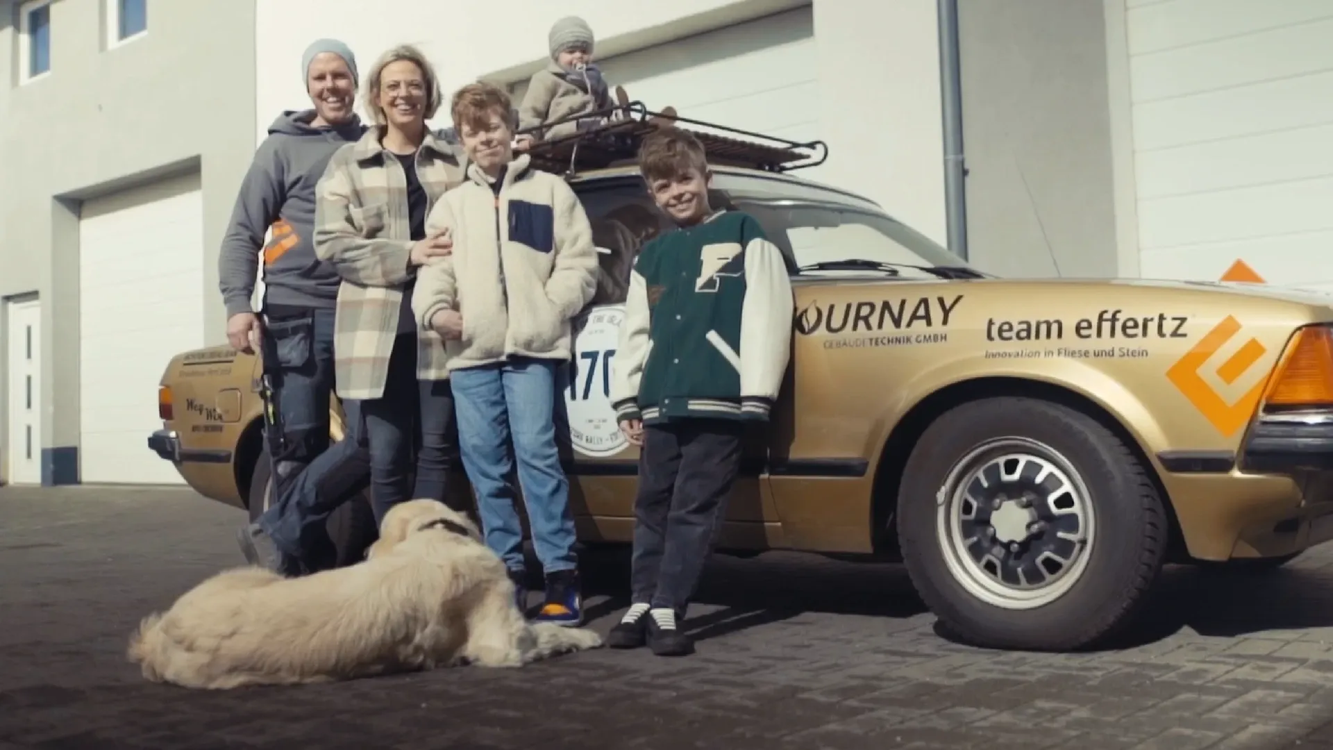 Fünf Personen vor einem goldenen Auto mit Werbeaufklebern; ein Kleinkind sitzt auf dem Dachgepäckträger, ein großer Hund liegt davor, im Hintergrund Garagentore