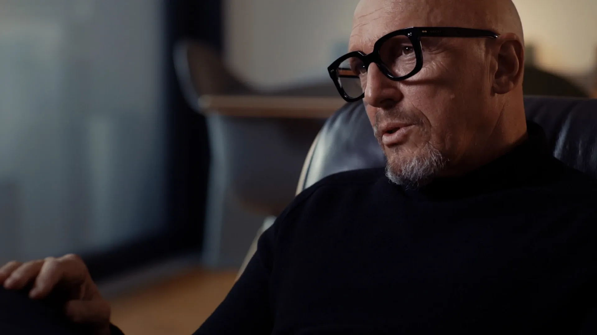 Glatzköpfiger Mann mit schwarzer Brille und Kinnbart in dunklem Pullover, seitliches Porträt im Innenraum